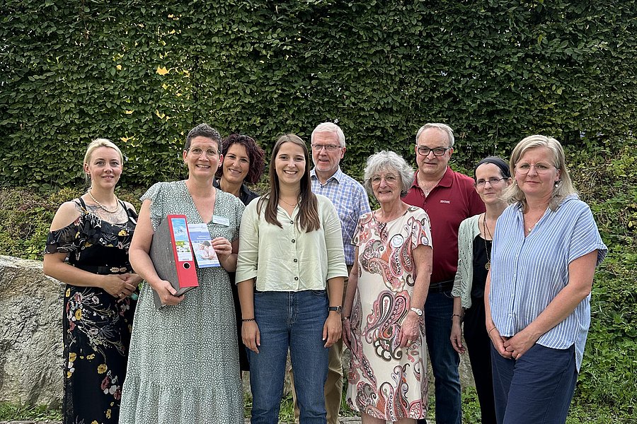 Neun Personen stehen in einem Garten vor einer hohen Hecke und einem Hausdach im Hintergrund