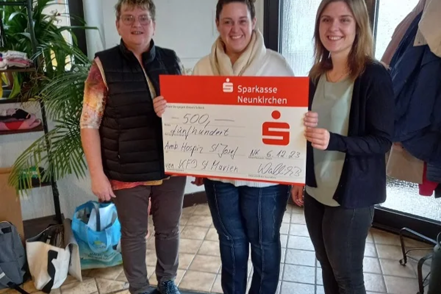 Drei Personen stehen in einem Raum mit Holzboden, eine hält einen großen roten Scheck der Sparkasse Neunkirchen über 500 Euro.