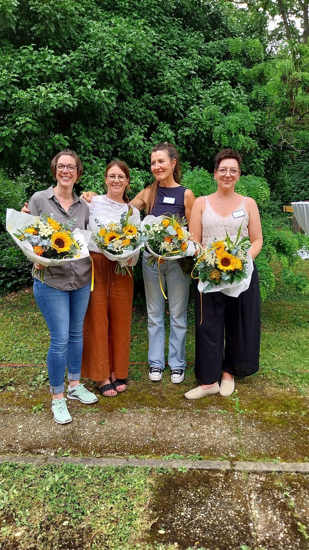 Vier Frauen stehen nebeneinander im Freien, jede hält einen Blumenstrauß mit Sonnenblumen, im Hintergrund dichter grüner Bewuchs