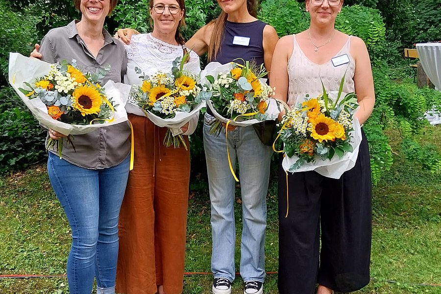 Vier Frauen stehen nebeneinander im Freien, jede hält einen Blumenstrauß mit Sonnenblumen, im Hintergrund dichter grüner Bewuchs