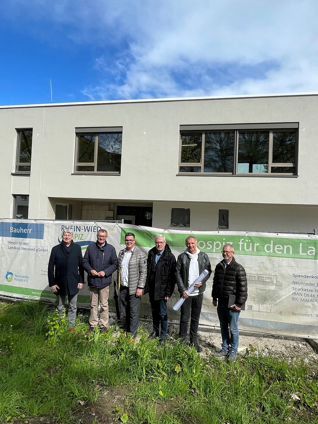 Sechs Personen stehen vor einer Baustelle mit Bauzaun und Werbebannern unter blauem Himmel
