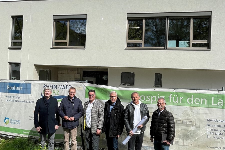 Sechs Personen stehen vor einer Baustelle mit Bauzaun und Werbebannern unter blauem Himmel