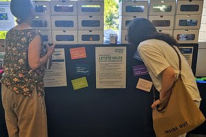 Zwei Frauen betrachten ein schwarzes Informationsboard mit mehreren kleinen Fächern und bunten Zetteln, im Hintergrund Fenster mit Bäumen.