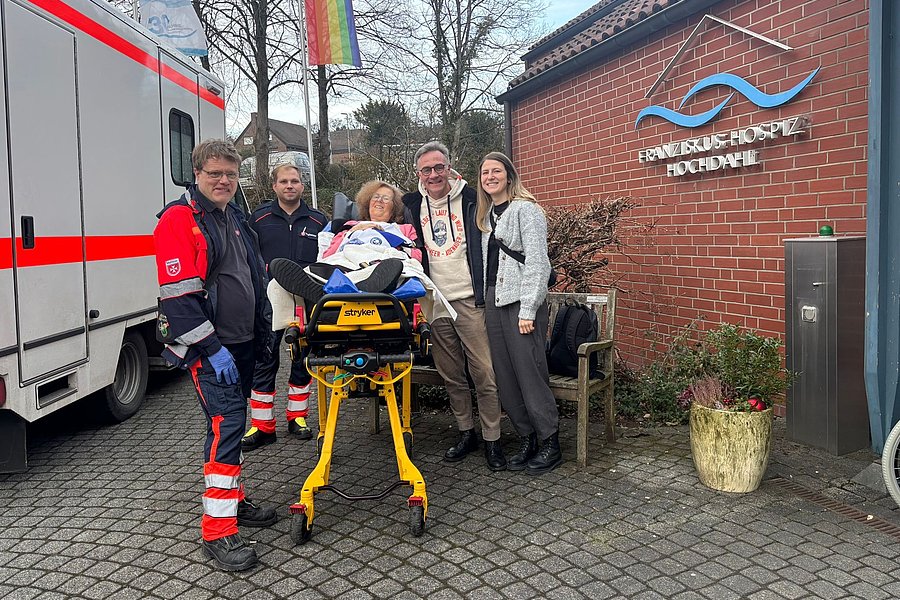 Gruppe von fünf Personen vor einem Gebäude, Rettungswagen im Hintergrund, helfende Hände für eine Person auf einer Trage.