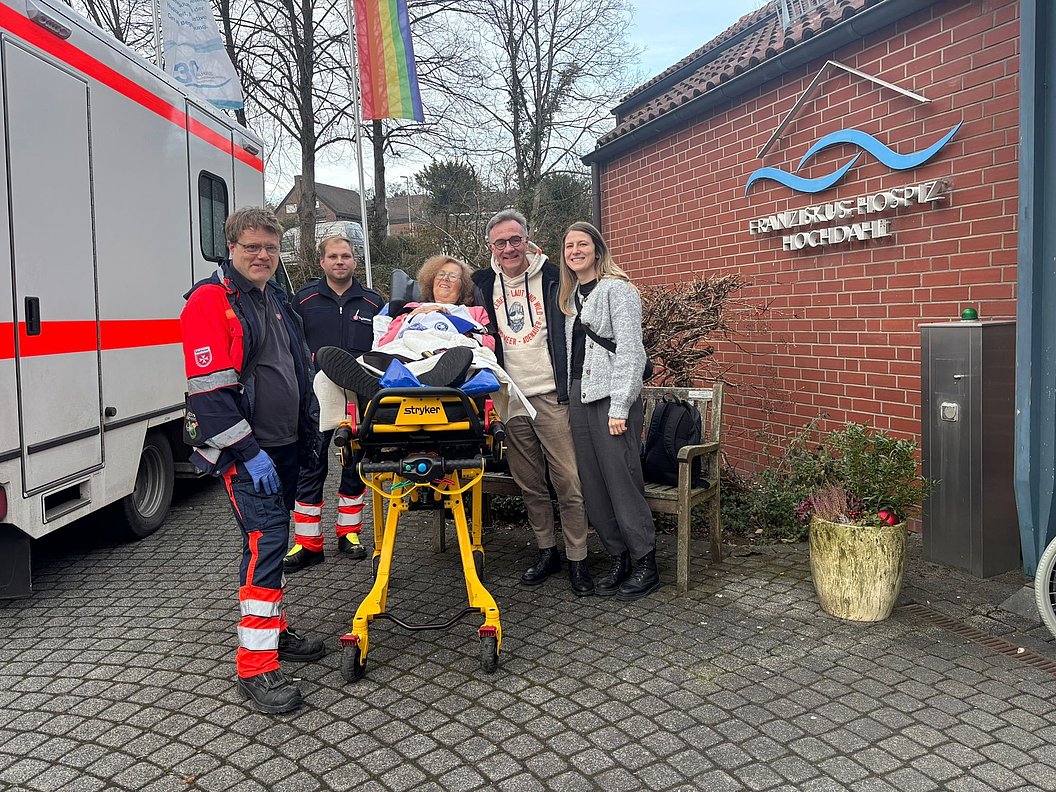 Gruppe von fünf Personen vor einem Gebäude, Rettungswagen im Hintergrund, helfende Hände für eine Person auf einer Trage.