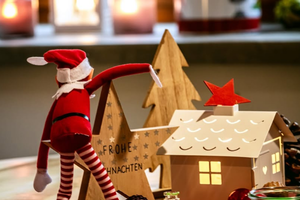 Weihnachtliche Szene mit einer kleinen Elf-Figur, die eine Papiertanne neben einem beleuchteten Papierhaus mit rotem Stern auf dem Dach bemalt, umgeben von mehreren kleinen Kerzengläsern auf einem roten Tuch.