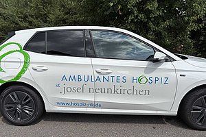 Weißer VW Polo mit der Aufschrift 'Ambulantes Hospiz st. josef neunkirchen' und der Website 'www.hospiz-nk.de' auf der Fahrzeugseite.