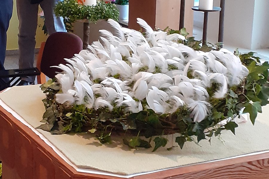 Kranz mit weißen Federn und grünen Blättern auf einem hölzernen Altar in einem Kirchenraum.