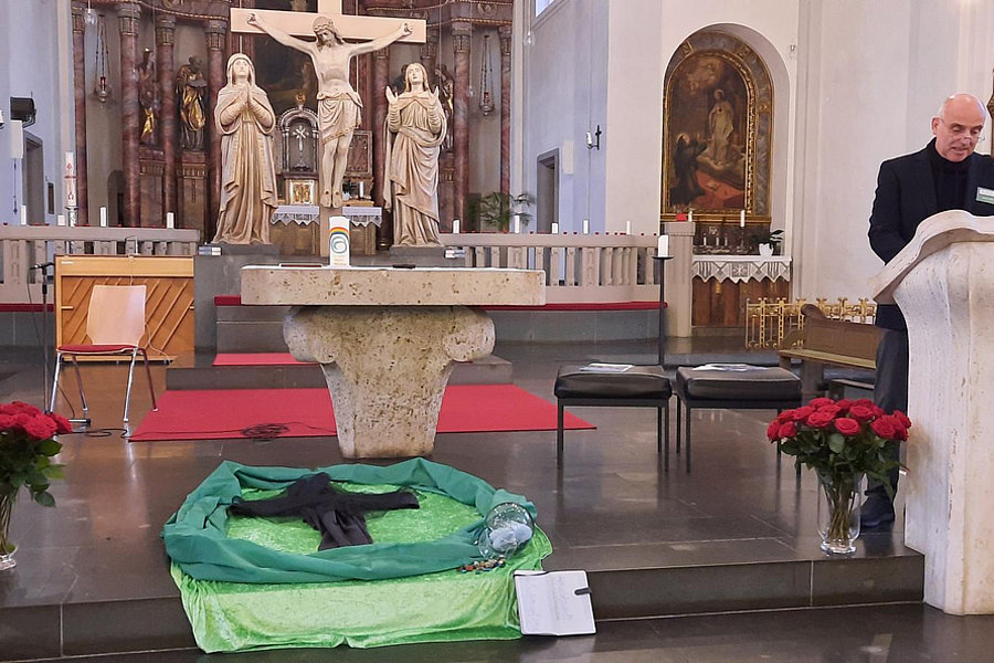 Kirchenschiff mit Altar, Kreuz, Statuen und mehreren Pflanzen, rechts ein Rednerpult mit einer Person.