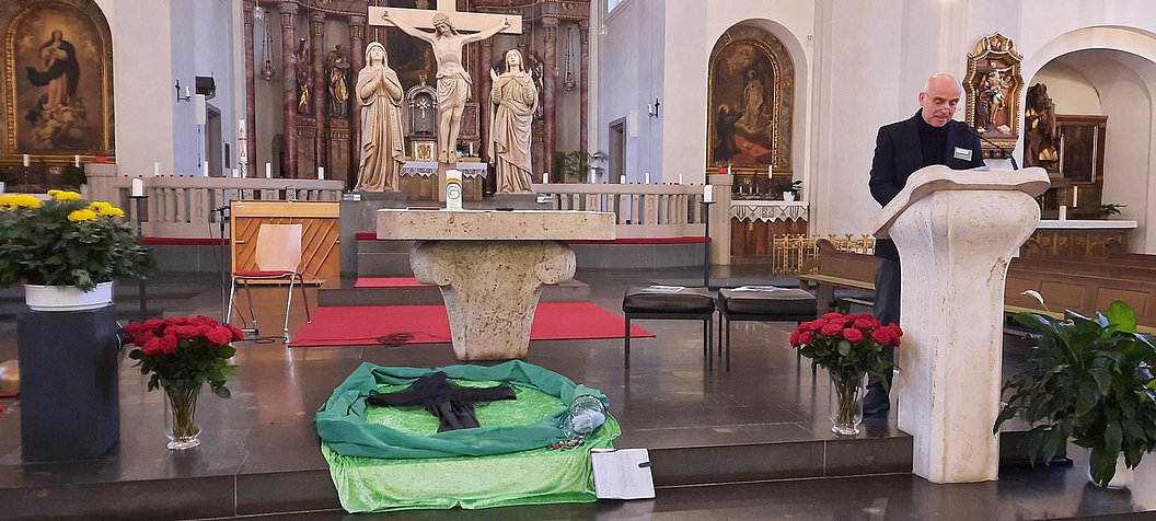 Kirchenschiff mit Altar, Kreuz, Statuen und mehreren Pflanzen, rechts ein Rednerpult mit einer Person.