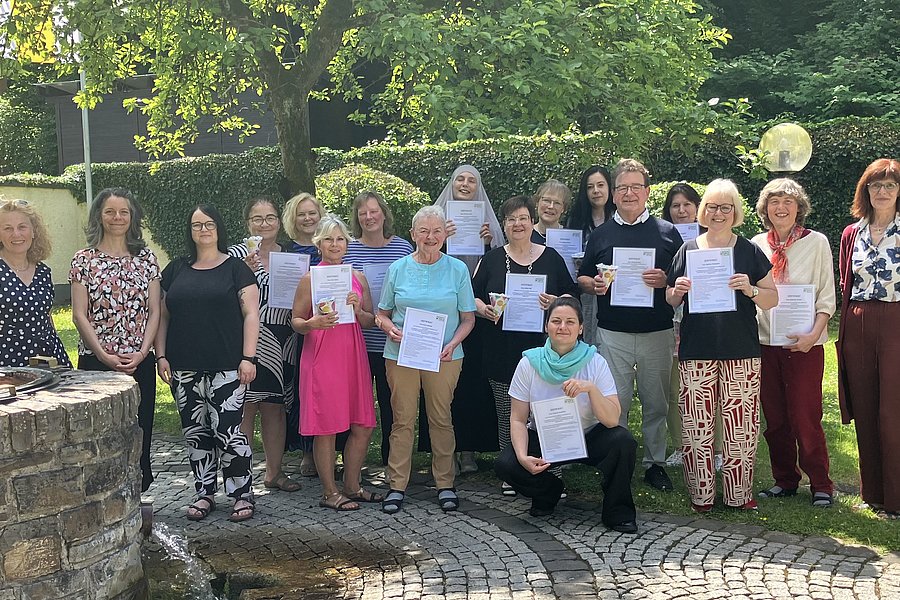 Gruppe von Erwachsenen steht im Garten, einige halten Urkunden in der Hand, umgeben von Bäumen und Hecken.