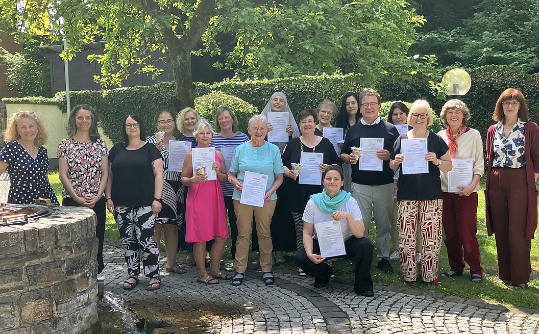 Gruppe von Erwachsenen steht im Garten, einige halten Urkunden in der Hand, umgeben von Bäumen und Hecken.