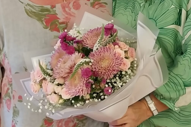 Zwei Frauen, eine mit grünem, gemustertem Oberteil und Namensschild, die andere mit hellgrauem Oberteil mit Blumenmuster, halten einen Blumenstrauß mit rosa Chrysanthemen und weißen Schleierkrautblüten.