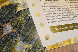 Ausschnitt einer Rezeptseite mit Text und dekorativem Blattmuster am linken Rand auf Holzuntergrund