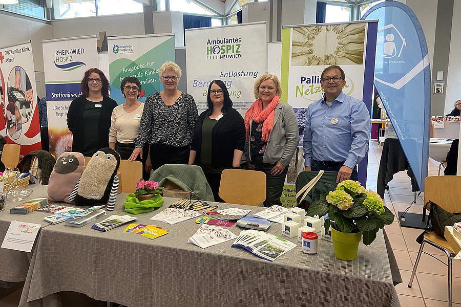 Sechs Personen stehen hinter einem Tisch mit Informationsmaterialien, zwei Plüschtiere in Schaf-Form liegen auf dem Tisch, im Hintergrund Banner mit Texten zu Hospiz und Palliativnetz.