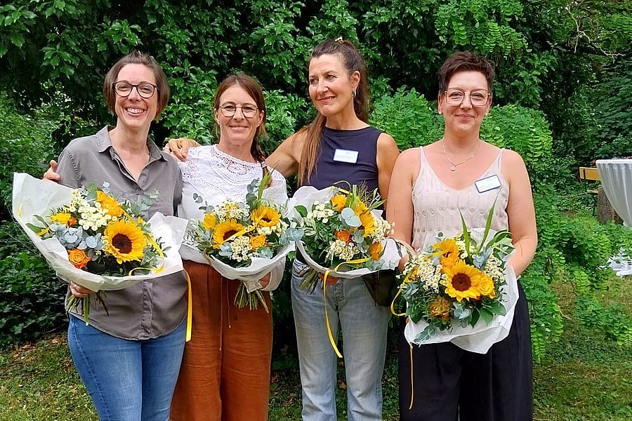 Vier Personen stehen nebeneinander im Freien, jede hält einen Blumenstrauß mit Sonnenblumen, umgeben von grünen Bäumen.
