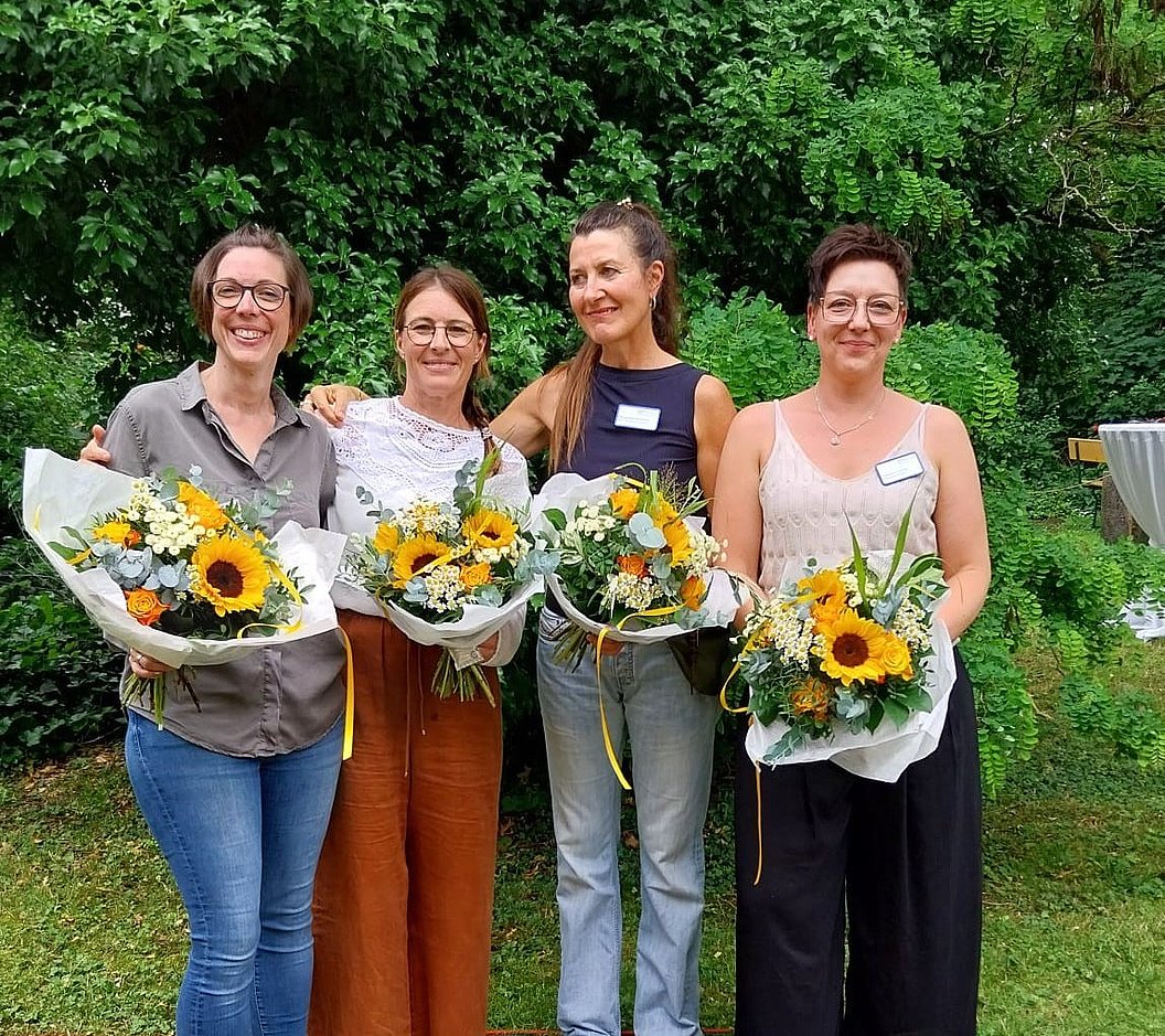 Vier Personen stehen nebeneinander im Freien, jede hält einen Blumenstrauß mit Sonnenblumen, umgeben von grünen Bäumen.