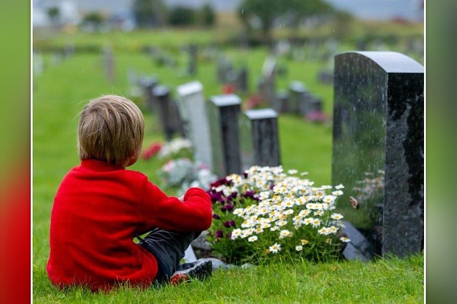 Kind sitzt auf einer Wiese vor einem Grabstein auf einem Friedhof, umgeben von Blumen.