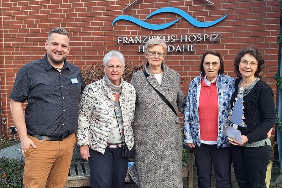 Fünf Personen stehen vor einer Backsteinwand mit dem Schriftzug 'FRANZISKUS-HOSPIZ HILDDAHL', zwei Frauen tragen gemusterte Jacken, eine Frau einen langen Mantel, ein Mann ein dunkles Hemd und eine weitere Frau einen schwarzen Pullover mit Schal.