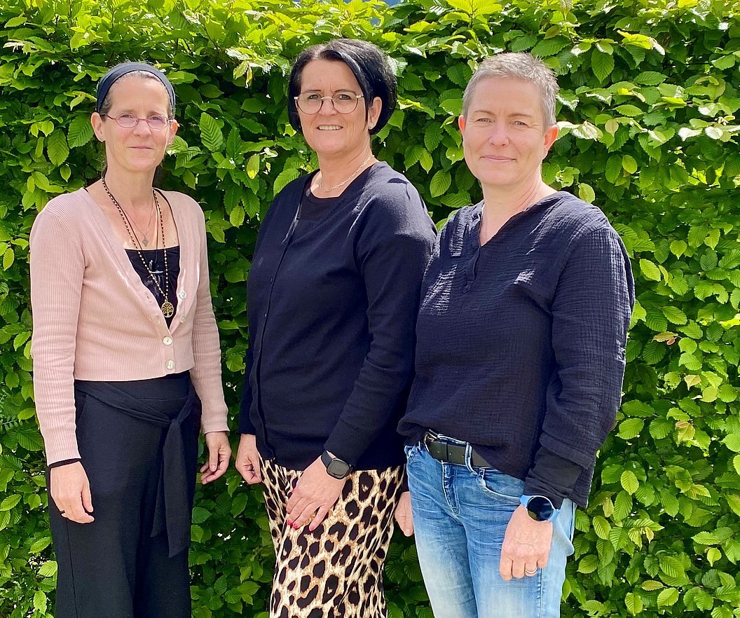 Drei Frauen stehen nebeneinander vor einer grünen Hecke, tragen unterschiedliche Kleidung: rosa Cardigan mit schwarzem Rock, schwarzes Oberteil mit Leopardenmuster-Rock, schwarzes Oberteil mit blauer Jeans.