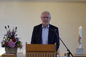 Mann in dunklem Anzug steht hinter einem hölzernen Rednerpult mit Mikrofon, links ein großer Blumenstrauß, rechts eine weiße Kerze auf einem Podest.