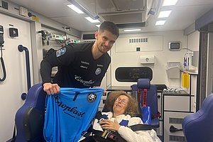 Person in Uniform hält ein blaues Trikot neben einer sitzenden Person mit Sicherheitsgurt in einem Rettungswagen