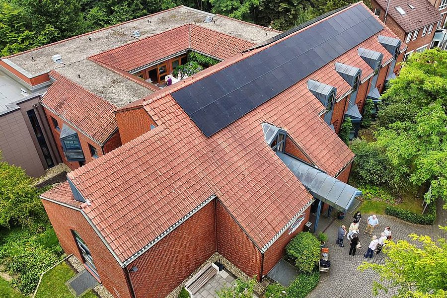 Luftaufnahme eines roten Backsteingebäudes mit Solarpaneelen auf dem Dach und mehreren Personen vor dem Eingang.