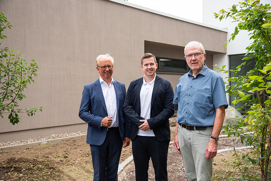 Drei Männer in Anzug und Hemd stehen vor einem modernen Gebäude mit brauner Fassade und begrünen Umgebung.