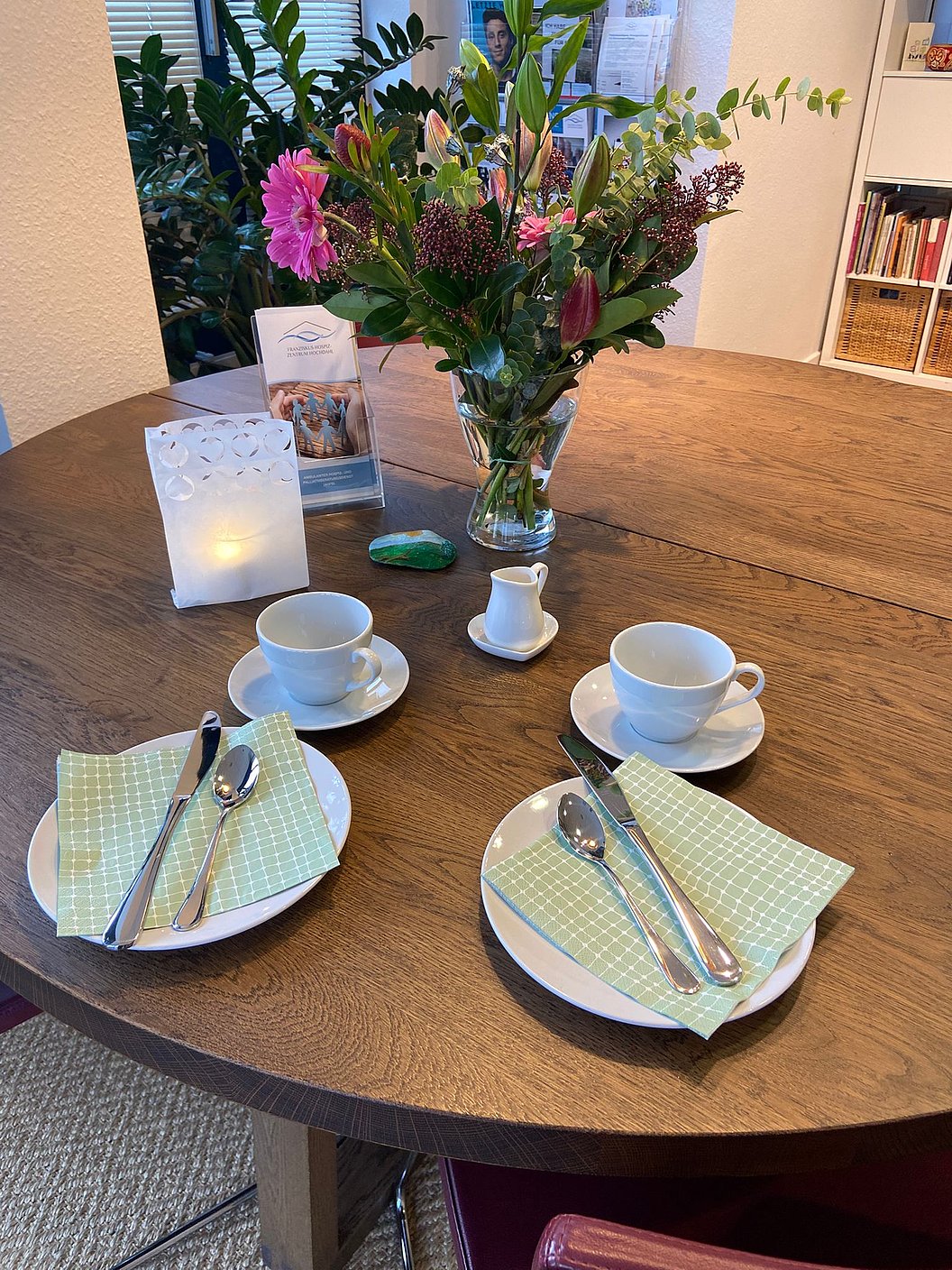Runder Holztisch mit zwei gedeckten Plätzen, grünen Servietten, Besteck, zwei Tassen, Milchgießer, Kerze und großem Blumenstrauß in Vase.