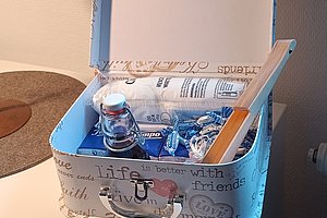 Offener, rechteckiger Koffer mit abgerundeten Ecken, bedruckt mit englischen Worten, gefüllt mit einer Glasflasche, einer Packung und mehreren kleinen blauen Gegenständen.