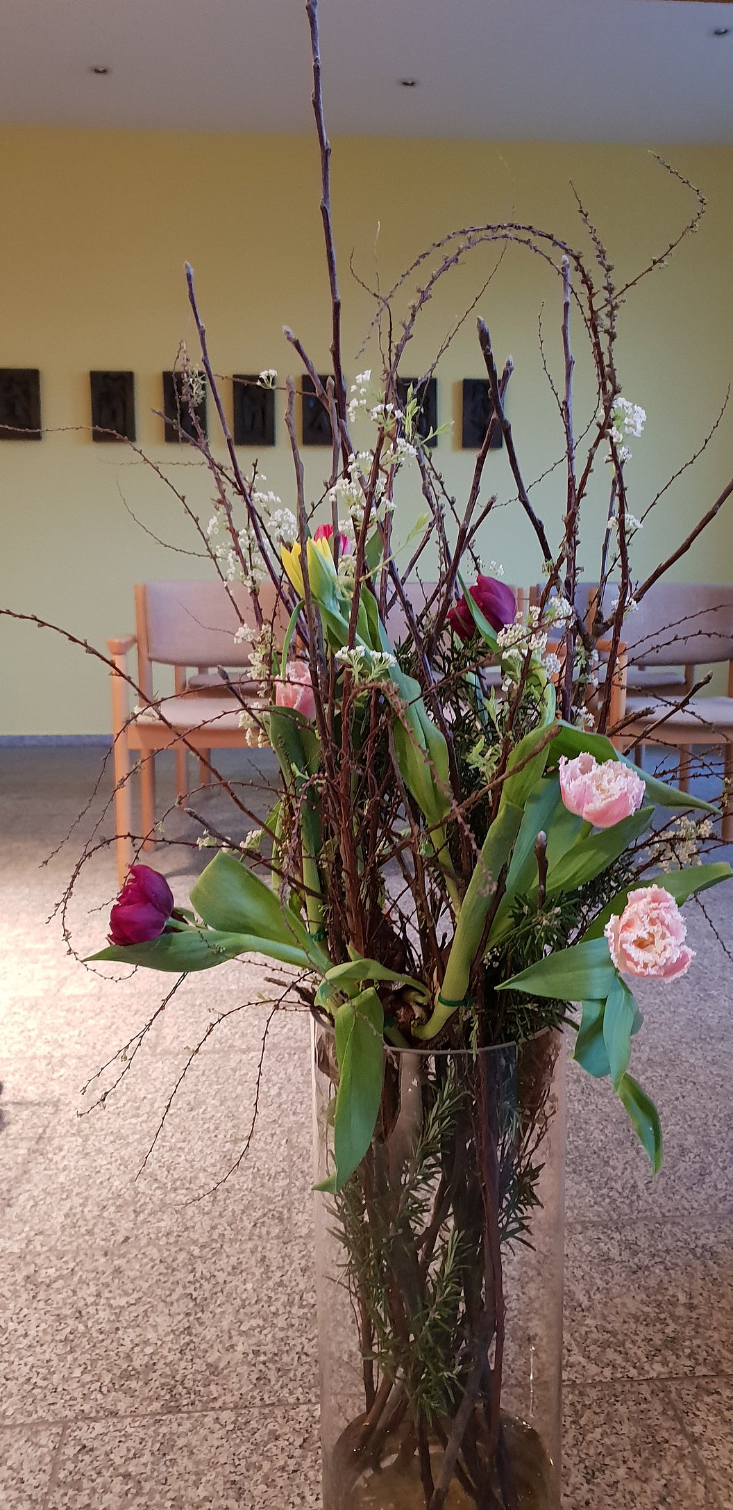 Blumengesteck mit Zweigen, grünen Blättern und rosa sowie roten Tulpen in einem hohen Glasvasenständer in einem Raum mit Stühlen und Bildern an der Wand