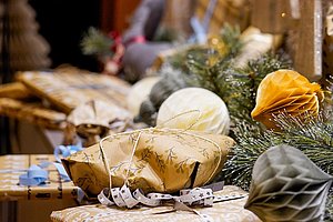 Mehrere festlich verpackte Geschenke mit Bändern liegen auf einem Tisch, daneben liegen Tannenzweige und dekorative Papierkugeln.