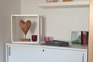 Weißer Schrank mit zwei Türen, darauf ein Holzherz in weißem Rahmen, zwei kleine rote Kerzenhalter, eine schwarze Schachtel und zwei Bücher. Darüber zwei weiße Wandregale mit einem Holzengel und einer weißen Kugel mit roten Mustern.