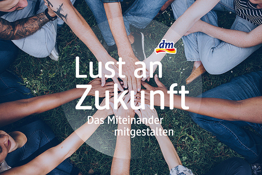 Mehrere Menschen sitzen im Kreis auf Gras und legen ihre Hände in die Mitte, darüber der Text 'Lust an Zukunft Das Miteinander mitgestalten' und das dm-Logo.