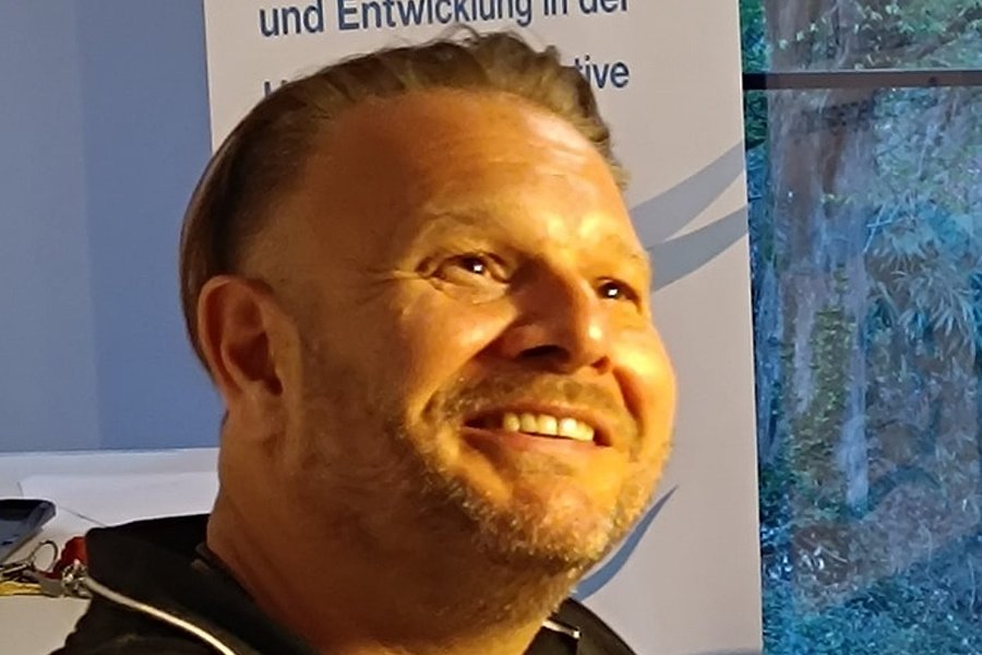 Mann mit dunklem Poloshirt vor Banner mit Text '30 Jahre Engagement, Kompetenz und Entwicklung in der ...' und Fenster mit Blick auf bewaldete Fläche.