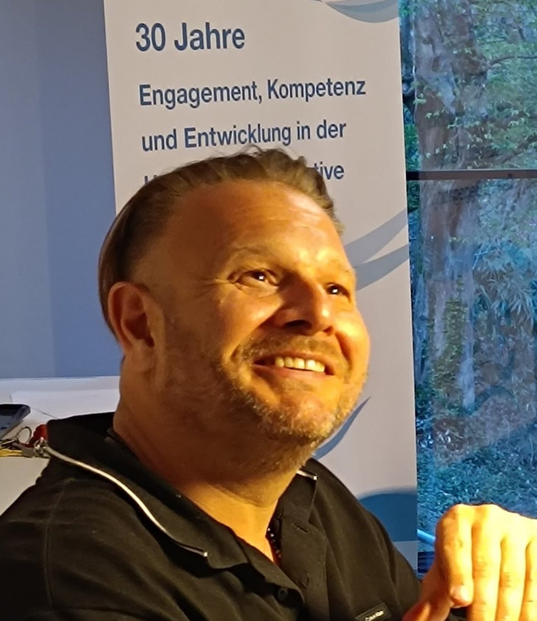 Mann mit dunklem Poloshirt vor Banner mit Text '30 Jahre Engagement, Kompetenz und Entwicklung in der ...' und Fenster mit Blick auf bewaldete Fläche.