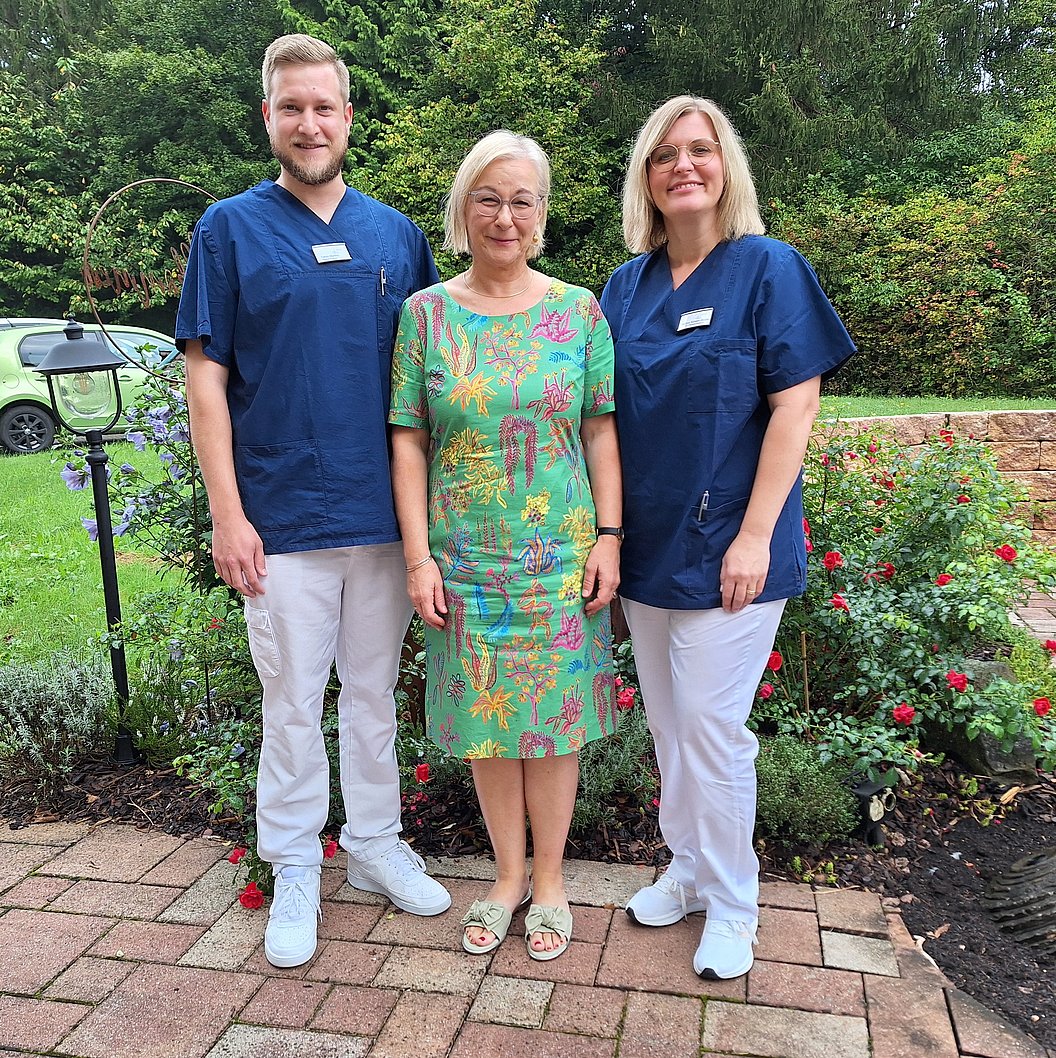 Drei Personen stehen nebeneinander auf einem gepflasterten Weg vor einem Garten mit Rosen und Büschen, zwei tragen blaue medizinische Oberteile und weiße Hosen, eine trägt ein grünes Kleid mit buntem Muster.
