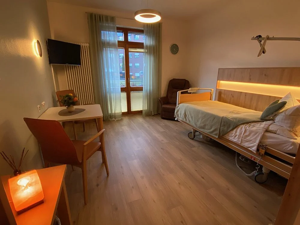 Krankenhauszimmer mit Pflegebett, Sessel, Tisch mit Stuhl, Fenster mit Gardinen und Fernseher an der Wand