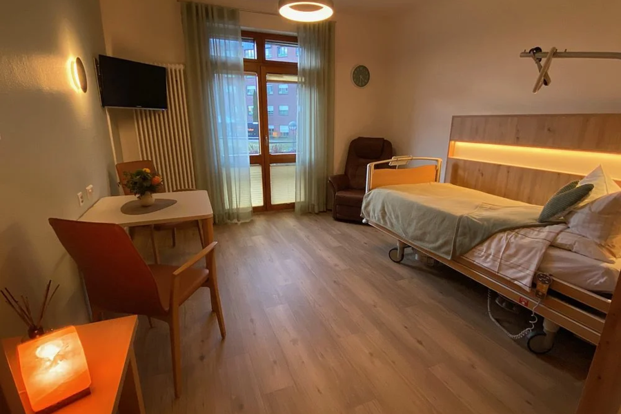Krankenhauszimmer mit Pflegebett, Sessel, Tisch mit Stuhl, Fenster mit Gardinen und Fernseher an der Wand