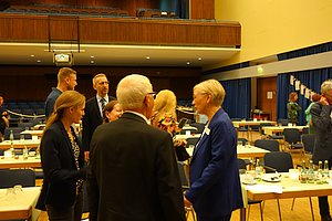 Mehrere Personen stehen in einem Saal mit Tischen, auf denen Flaschen und Gläser stehen, und unterhalten sich.