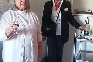 Zwei Frauen in einem Raum, eine hält ein Glas mit Getränk, die andere steht neben einem Servierwagen mit Flasche und Gläsern.