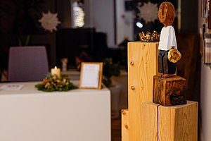 Holzskulptur einer stehenden Figur mit Hut auf einer mehrstufigen Holzsäule in einem Raum mit weihnachtlicher Dekoration.