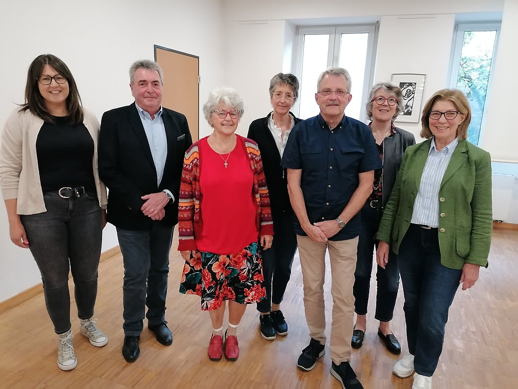 Gruppe von sieben Personen in einem hellen Raum, unterschiedliche Kleidung, stehend, einige tragen Jacken oder Blazer.