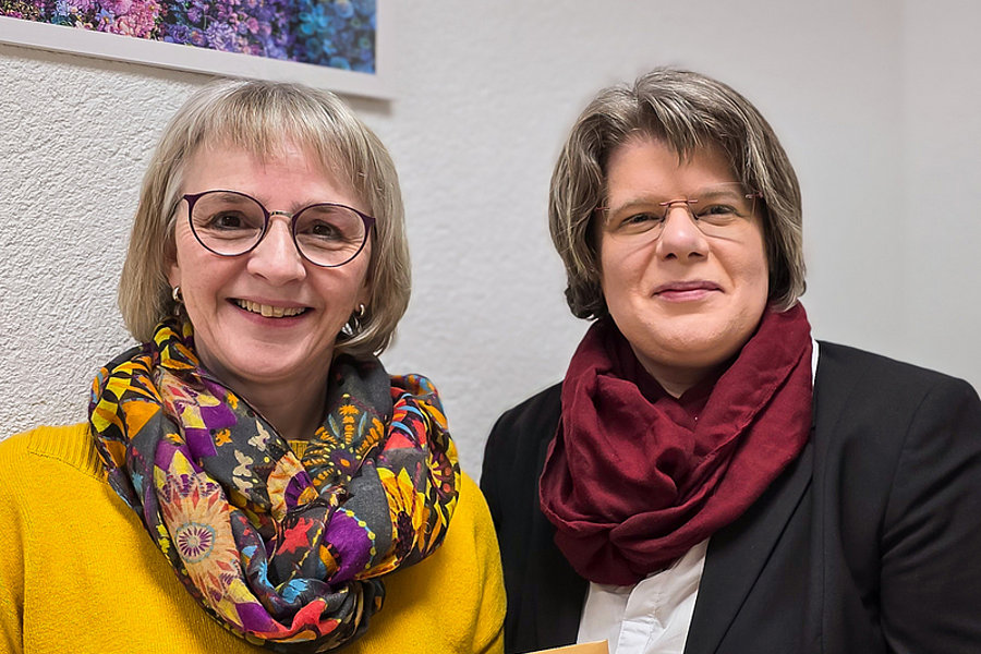 Zwei Frauen sitzen nebeneinander, eine trägt einen gelben Pullover und einen bunten Schal, die andere einen schwarzen Blazer und einen roten Schal. Eine hält einen braunen Umschlag mit handschriftlichem Text.