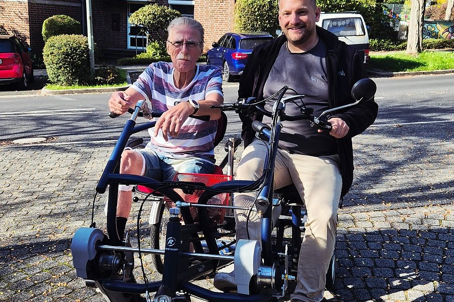 Zwei Personen sitzen auf einem dreirädrigen Tandemfahrrad auf einer gepflasterten Straße vor einem Backsteingebäude.