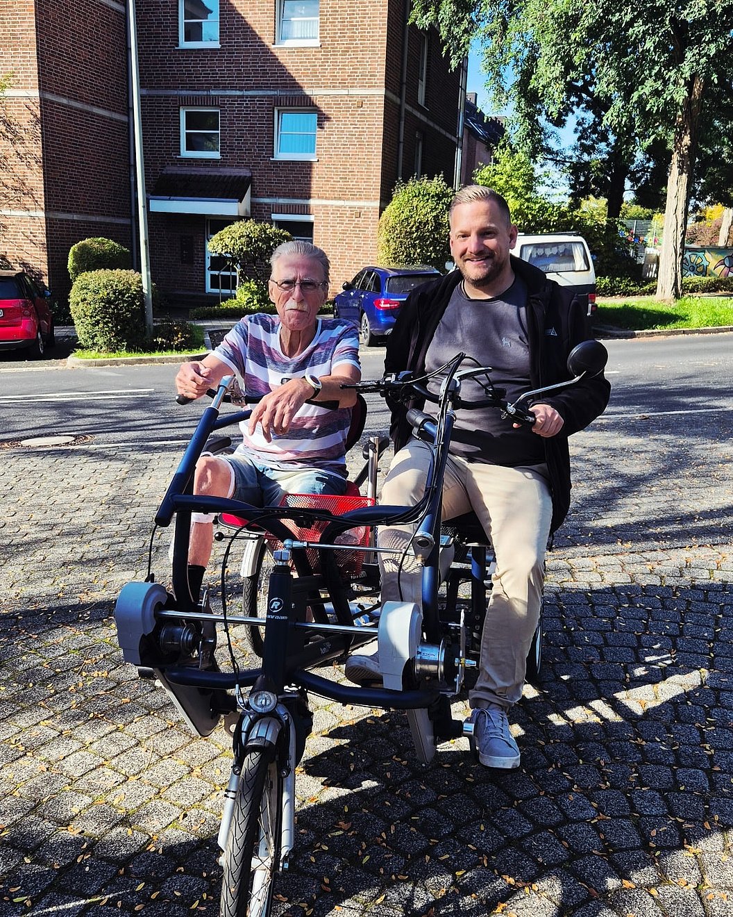 Zwei Personen sitzen auf einem dreirädrigen Tandemfahrrad auf einer gepflasterten Straße vor einem Backsteingebäude.