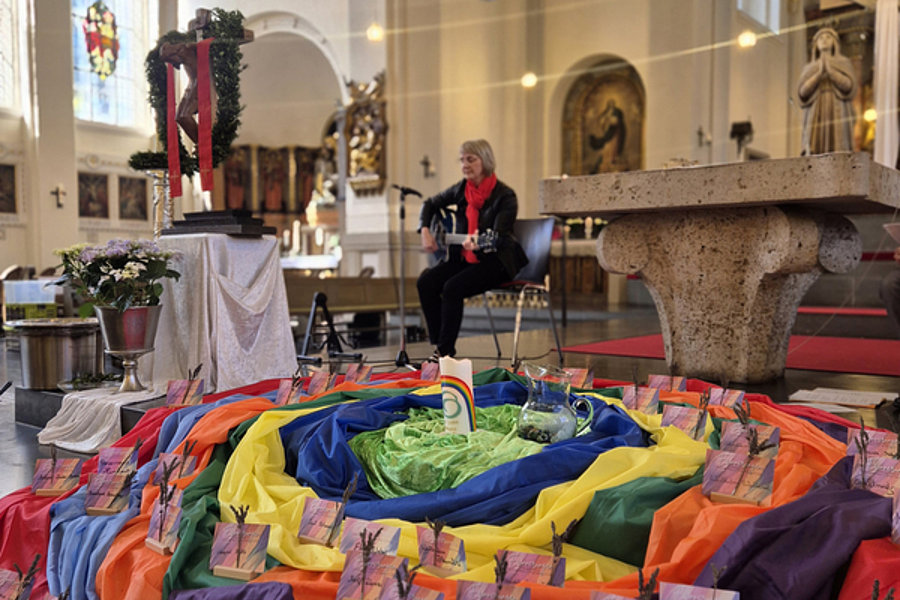 Bunte Stoffbahnen in Spirale auf dem Boden einer Kirche, darauf kleine Karten mit Windmühlenmotiven, im Hintergrund eine Person mit Akkordeon vor Altar und Kirchenfenstern.