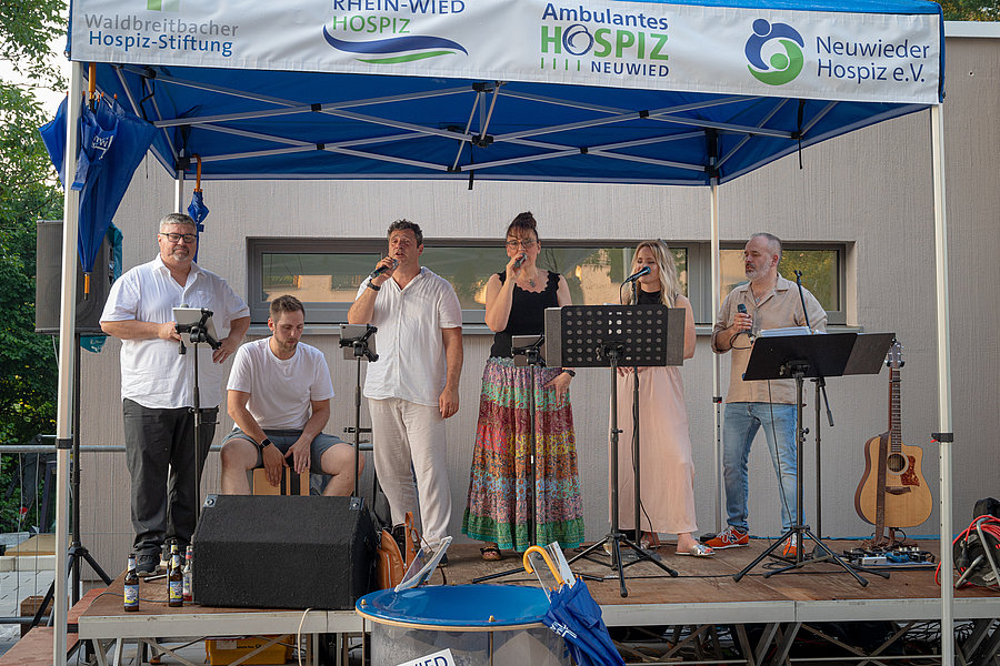 Sechs Personen auf einer Bühne unter einem blauen Zelt mit Bannern von Hospiz-Stiftungen, sie singen und spielen Musik, eine Gitarre steht rechts