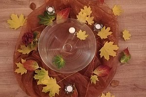 Herbstliche Dekoration mit gelben und roten Blättern, drei kleinen weißen Kerzen und einer großen Glasvase auf einem Holzboden