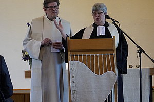 Zwei Geistliche in liturgischer Kleidung stehen an einem hölzernen Lesepult mit weißem Tuch, Mikrofon daneben.